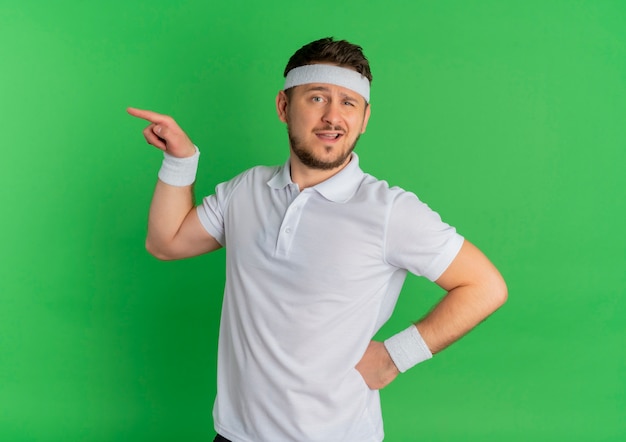 Hombre joven fitness en camisa blanca con diadema mirando hacia el frente apuntando con el dedo hacia el lado de pie sobre la pared verde