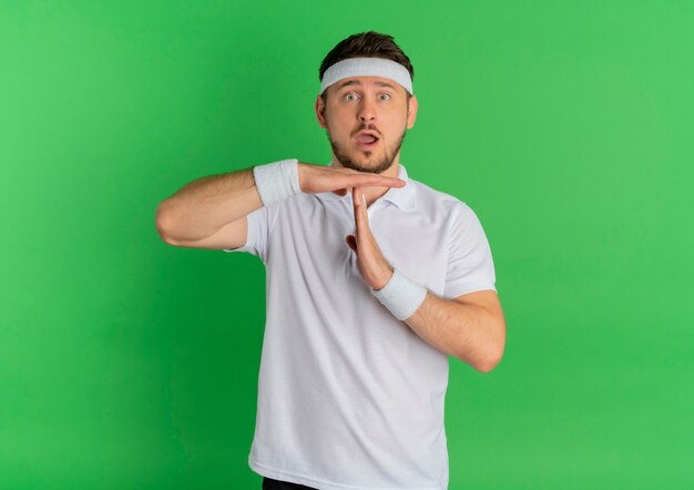 Hombre joven fitness en camisa blanca con diadema mirando hacia el frente haciendo gesto de tiempo de espera con las manos sorprendidas de pie sobre la pared verde