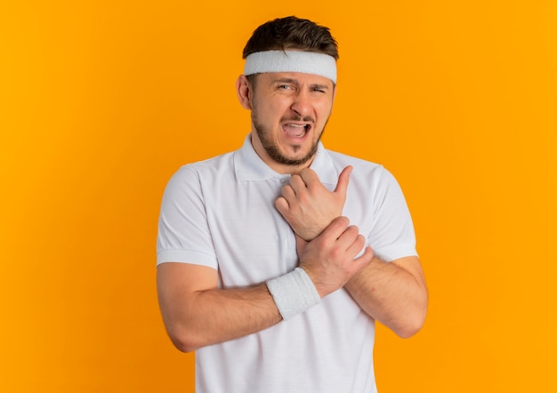 Hombre joven fitness en camisa blanca con diadema mirando hacia el frente tocando la muñeca con dolor de pie sobre la pared naranja