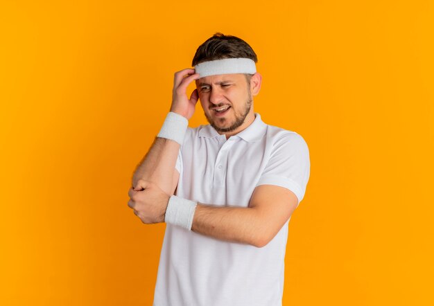 Hombre joven fitness en camisa blanca con diadema mirando a un lado confundido parado sobre fondo naranja