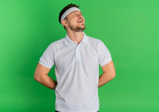 Hombre joven fitness en camisa blanca con diadema mirando a un lado con una sonrisa en la cara de pie sobre fondo verde