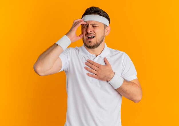 Hombre joven fitness en camisa blanca con diadema que parece mal con fuerte dolor de cabeza de pie sobre la pared naranja