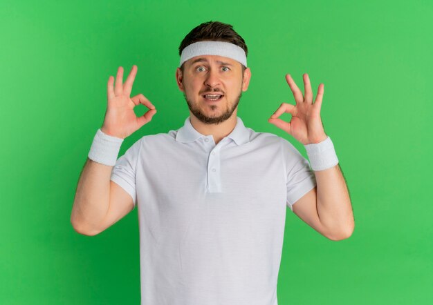 Hombre joven fitness en camisa blanca con diadema sonriendo haciendo bien firmar con ambas manos de pie sobre la pared verde