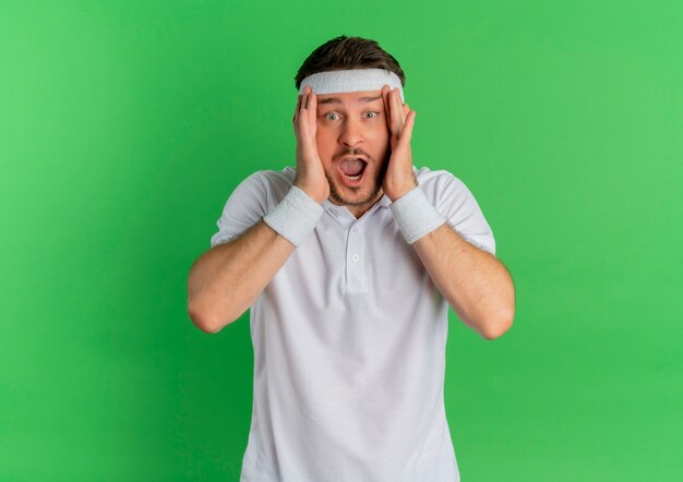 Hombre joven fitness en camisa blanca con diadema sorprendido sosteniendo su cabeza de pie sobre la pared verde