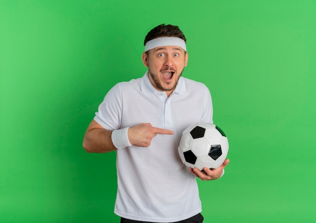 Hombre joven fitness en camisa blanca con diadema sosteniendo un balón de fútbol apuntando con el dedo índice a él de pie feliz y emocionado sobre fondo verde