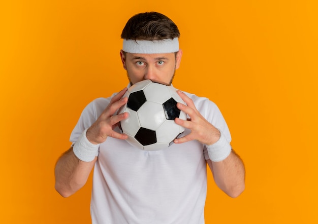 Hombre joven fitness en camisa blanca con diadema sosteniendo un balón de fútbol escondiendo la cara detrás de él de pie sobre fondo naranja