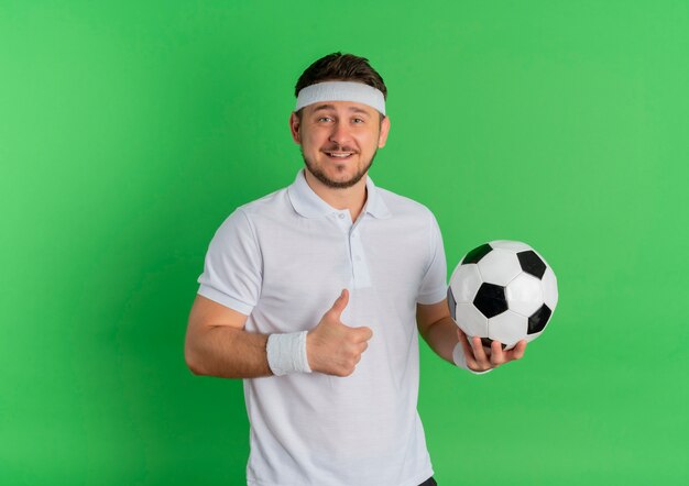 Hombre joven fitness en camisa blanca con diadema sosteniendo un balón de fútbol sonriendo alegremente mostrando los pulgares para arriba sobre fondo verde