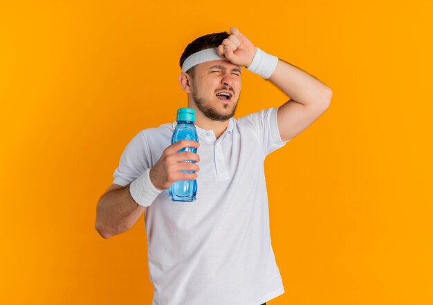 Hombre joven fitness en camisa blanca con diadema sosteniendo una botella de agua con aspecto cansado y agotado después del entrenamiento de pie sobre fondo naranja