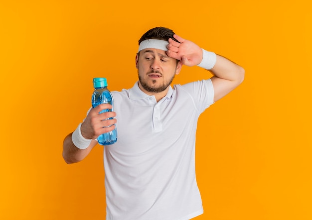 Hombre joven fitness en camisa blanca con diadema sosteniendo una botella de agua con aspecto cansado y agotado de pie sobre fondo naranja