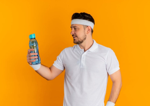 Hombre joven fitness en camisa blanca con diadema sosteniendo una botella de agua mirándolo de pie sobre fondo naranja