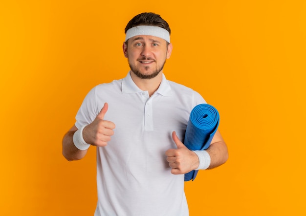 Hombre joven fitness en camisa blanca con diadema sosteniendo estera de yoga mostrando los pulgares para arriba mirando a la cámara sonriendo de pie sobre fondo naranja