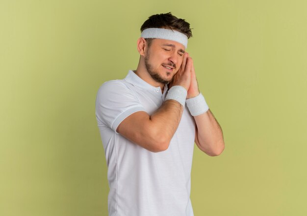 Hombre joven fitness en camisa blanca con diadema sosteniendo las palmas juntas apoyando la cabeza en las palmas con los ojos cerrados quiere dormir de pie sobre fondo verde oliva