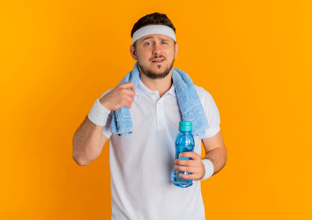 Hombre joven fitness en camisa blanca con diadema y toalla alrededor del cuello sosteniendo una botella de agua mirando a la cámara con expresión segura de pie sobre fondo naranja