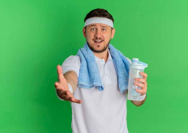 Hombre joven fitness en camisa blanca con diadema y toalla alrededor de su cuello sosteniendo una botella de agua mirando a la cámara sonriendo con el brazo extendido de pie sobre fondo verde