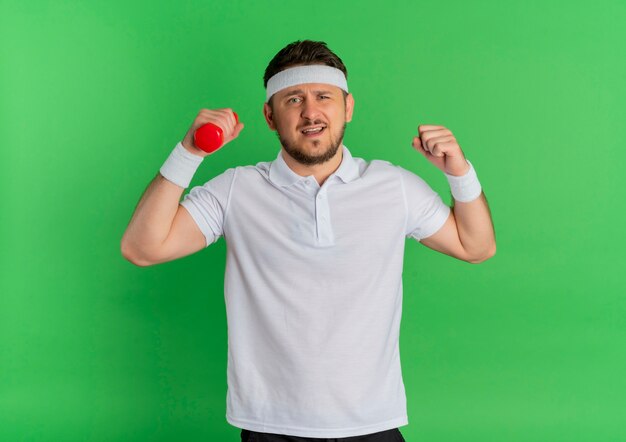 Hombre joven fitness en camisa blanca con diadema trabajando con mancuernas tensas y confiadas de pie sobre la pared verde