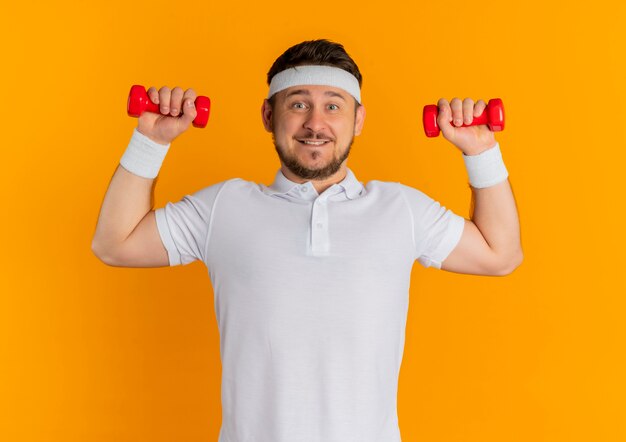 Hombre joven fitness en camisa blanca con diadema trabajando con pesas mirando confiado sonriendo de pie sobre la pared naranja