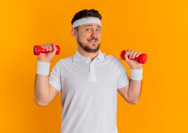 Hombre joven fitness en camisa blanca con diadema trabajando con pesas mirando confiado sonriendo de pie sobre la pared naranja