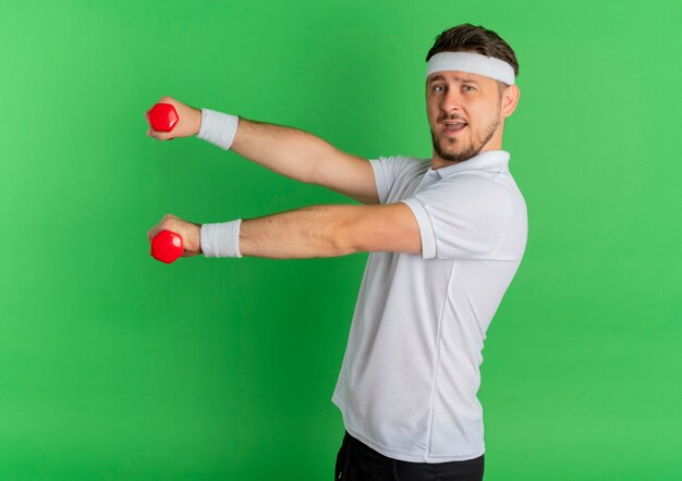 Hombre joven fitness en camisa blanca con diadema trabajando con pesas de pie feliz y positivo sobre pared verde