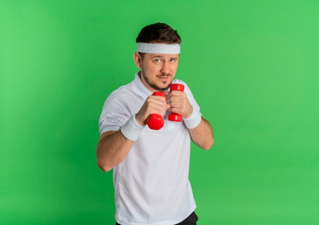 Hombre joven fitness en camisa blanca con diadema trabajando con pesas tensas de pie sobre fondo verde