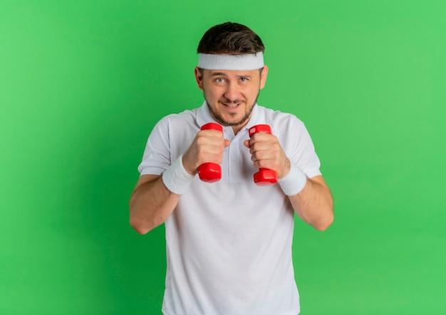 Hombre joven fitness en camisa blanca con diadema trabajando con pesas tensas de pie sobre la pared verde