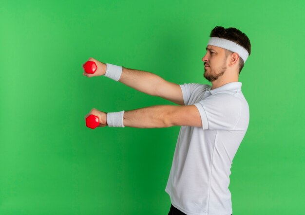 Hombre joven fitness en camisa blanca con diadema trabajando con pesas tensas de pie sobre la pared verde