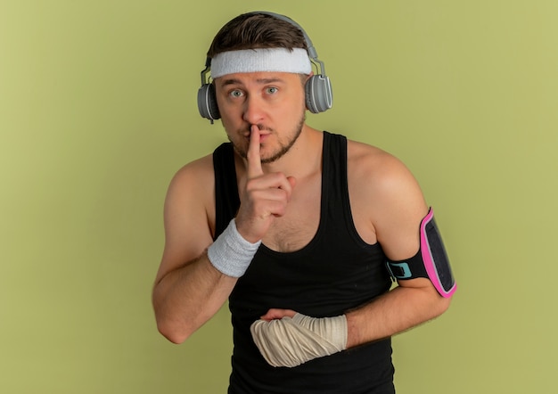 Hombre joven fitness con diadema y auriculares mirando a la cámara de entrenamiento con brazalete de teléfono inteligente haciendo gesto de silencio con el dedo en los labios sobre fondo verde oliva