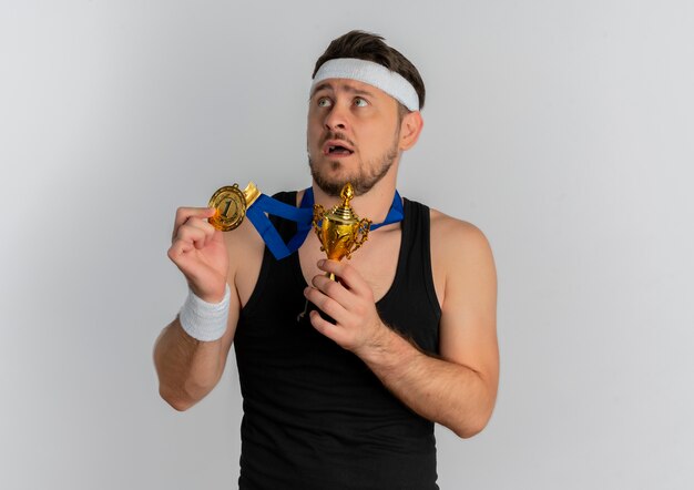 Hombre joven fitness con diadema y medalla de oro alrededor de su cuello sosteniendo su trofeo mirando a un lado con expresión de miedo de pie sobre fondo blanco