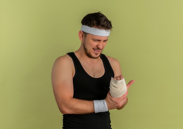 Hombre joven fitness con diadema mirando su muñeca vendada sintiendo dolor de pie sobre fondo verde oliva