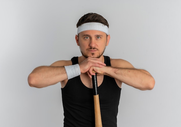 Hombre joven fitness con diadema sosteniendo un bate de béisbol mirando a la cámara con rostro serio de pie sobre fondo blanco.