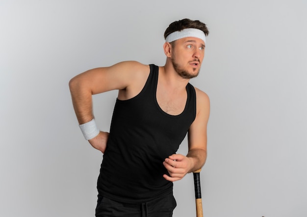 Hombre joven fitness con diadema sosteniendo un bate de béisbol mirando a un lado con expresión confusa de pie sobre fondo blanco.