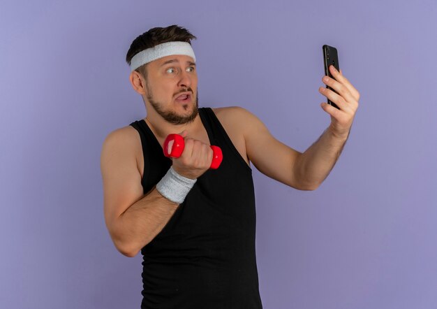 Hombre joven fitness con diadema tomando selfie usando su teléfono inteligente posando con mancuernas en la mano mirando confundido parado sobre pared púrpura