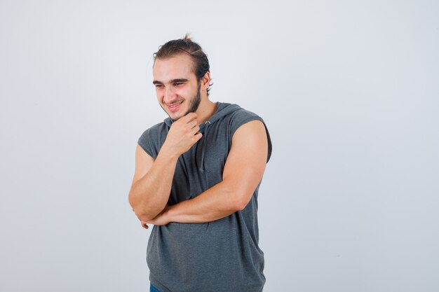 Hombre joven en forma manteniendo la mano debajo de la barbilla en una sudadera con capucha sin mangas y luciendo dichoso, vista frontal.