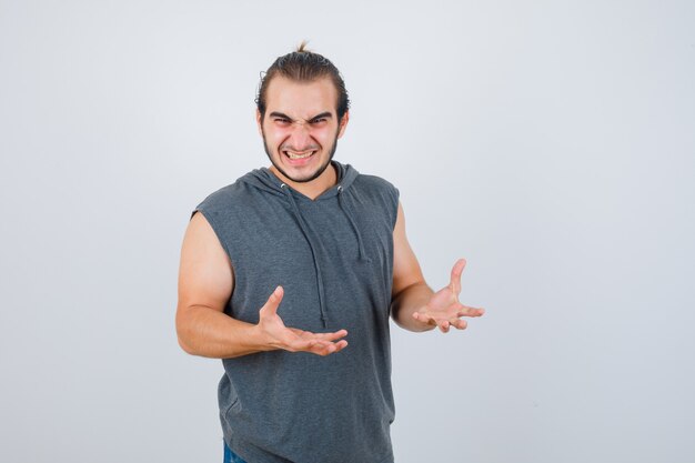 Hombre joven en forma manteniendo las manos de manera agresiva en una sudadera con capucha sin mangas y con aspecto ansioso. vista frontal.