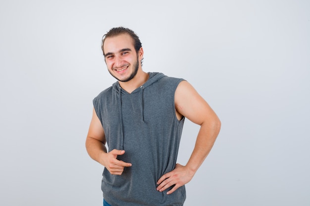 Hombre joven en forma posando con la mano en la cintura en una sudadera con capucha sin mangas y luciendo alegre. vista frontal.