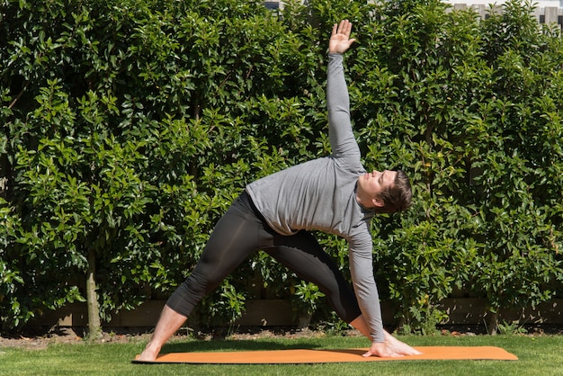 Foto gratuita hombre joven en forma practicando yoga y haciendo la pose de triángulo al aire libre