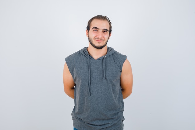 Hombre joven en forma con sudadera con capucha sin mangas manteniendo las manos detrás de la espalda y mirando alegre, vista frontal.