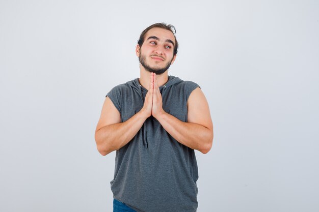 Hombre joven en forma con sudadera con capucha sin mangas manteniendo las manos en gesto de oración y mirando esperanzado, vista frontal.