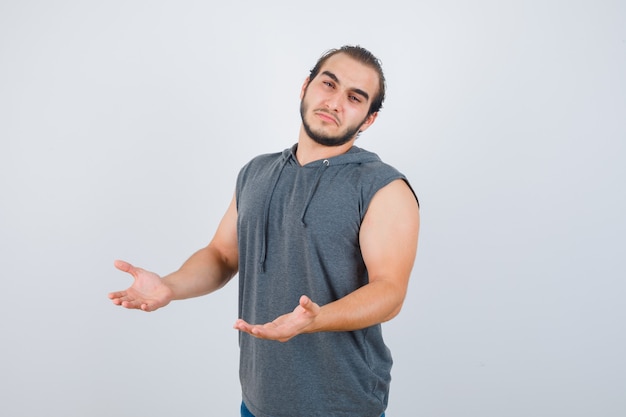 Hombre joven en forma con sudadera con capucha sin mangas que muestra un gesto de impotencia y parece molesto, vista frontal.