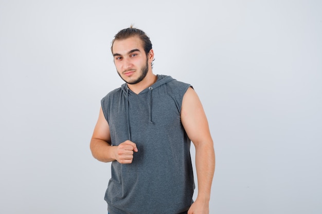 Hombre joven en forma con sudadera con capucha sin mangas que muestra el puño cerrado y parece seguro, vista frontal.