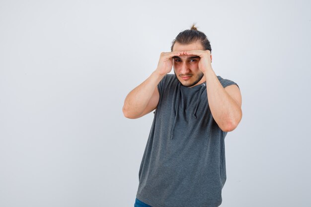 Hombre joven en forma con sudadera con capucha sin mangas sosteniendo las manos por encima de la cabeza para ver con claridad y mirando enfocado, vista frontal.