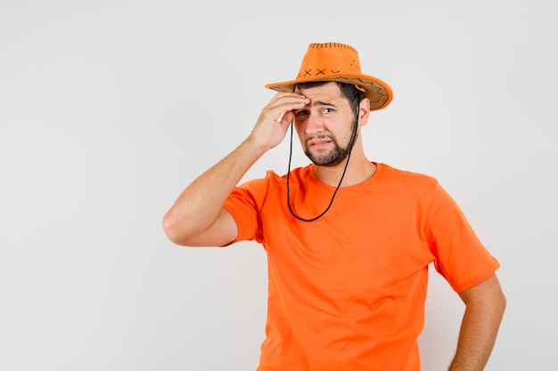 Hombre joven frotándose la frente en camiseta naranja, sombrero y mirando triste. vista frontal.