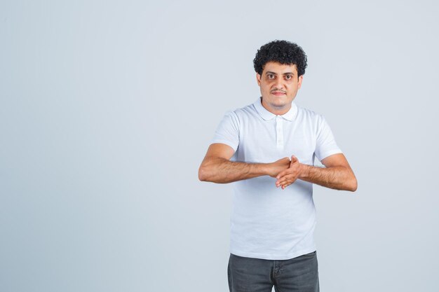 Hombre joven frotándose las manos en camiseta blanca y jeans y mirando serio, vista frontal.