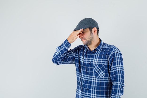 Hombre joven frotándose los ojos y la nariz en camisa, gorra y con aspecto cansado. vista frontal.