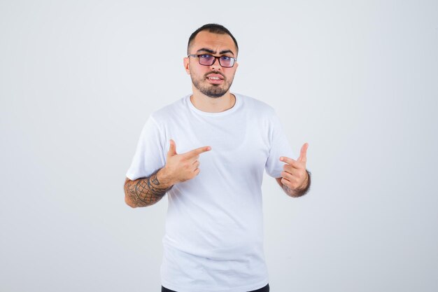 Hombre joven con gafas y apuntando a sí mismo con camiseta blanca y pantalón negro y mirando serio