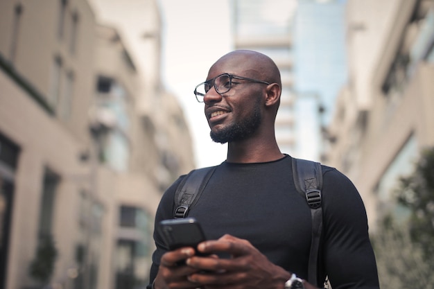 Hombre joven con gafas y una mochila sosteniendo un teléfono celular bajo la luz del sol