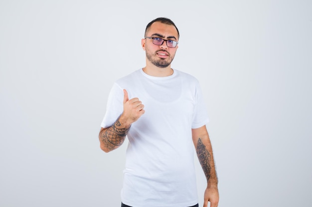 Hombre joven con gafas y mostrando el pulgar hacia arriba en camiseta blanca y pantalón negro y mirando feliz