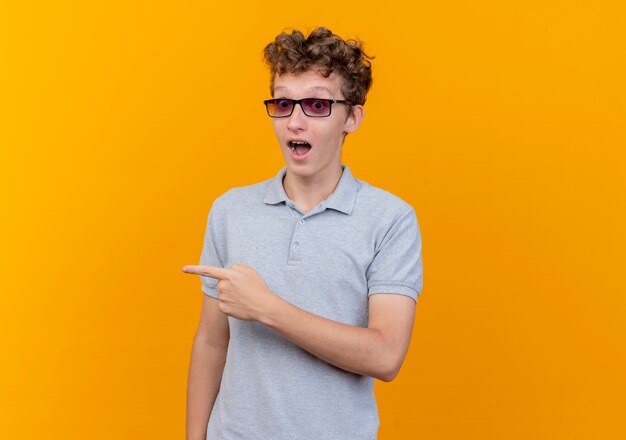 Hombre joven con gafas negras con camisa de polo gris asombrado y sorprendido apuntando con el índice figner a algo parado sobre la pared naranja