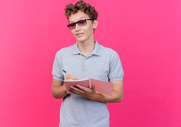 Hombre joven con gafas negras con camisa de polo gris con cuaderno con lápiz feliz y positivo sonriendo sobre rosa
