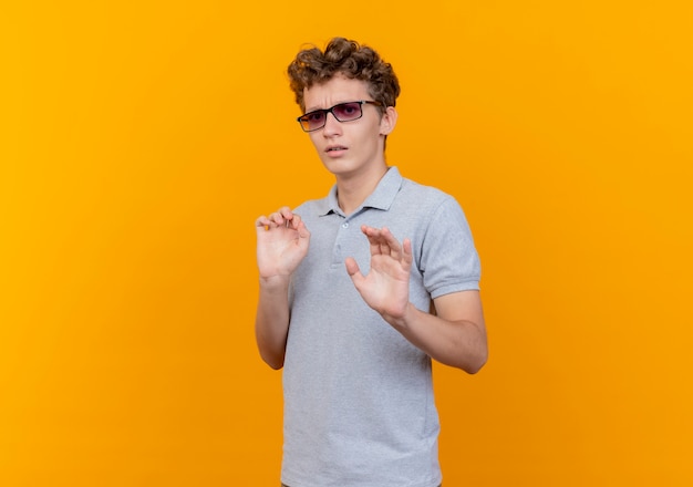 Hombre joven con gafas negras con camisa de polo gris haciendo señal de stop sosteniendo las manos como diciendo no te acerques asustado de pie sobre la pared naranja