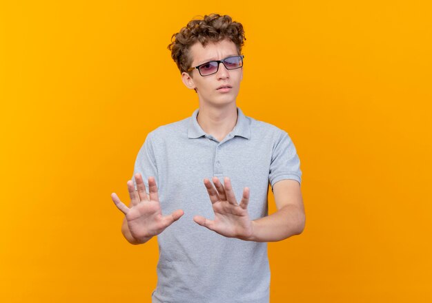 Hombre joven con gafas negras con camisa polo gris haciendo señal de stop sosteniendo las manos como diciendo no te acerques parado sobre la pared naranja